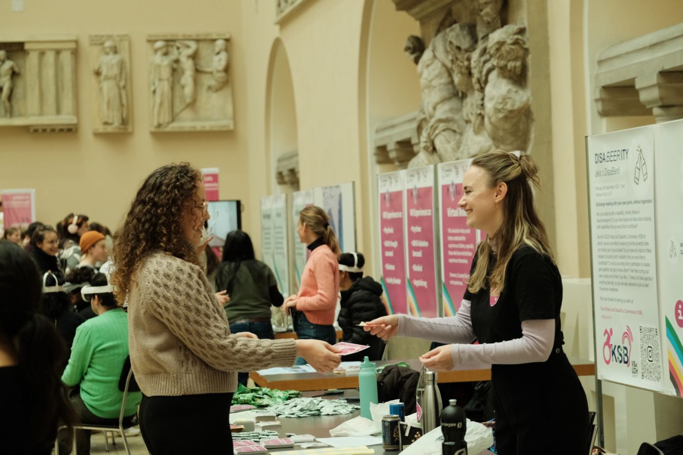Infostand KSB/VSUZH: Zwei Personen stehen sich an einem Infostand gegenüber und reichen sich Informationsmaterial, im Hintergrund weitere Stände und Besucher*innen.